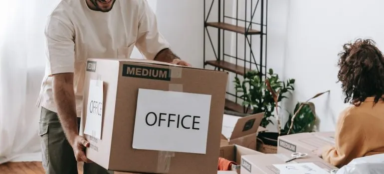 a man carrying a cardboard box that says "Office" while a woman packs in the background